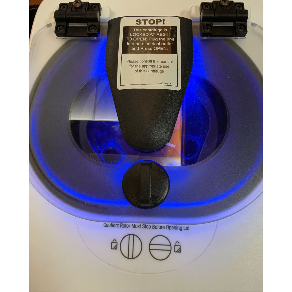 Close-up top view of the centrifuge's clear lid displaying a "STOP!" warning label, with bright blue LED lighting illuminating the internal chamber.   