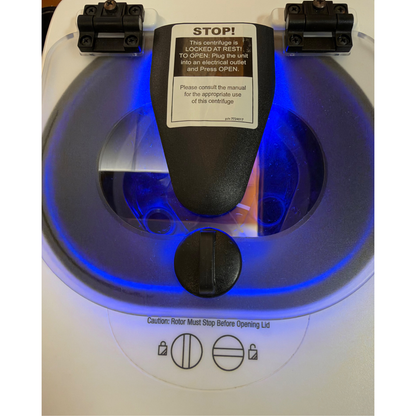 Close-up top view of the centrifuge's clear lid displaying a "STOP!" warning label, with bright blue LED lighting illuminating the internal chamber.   