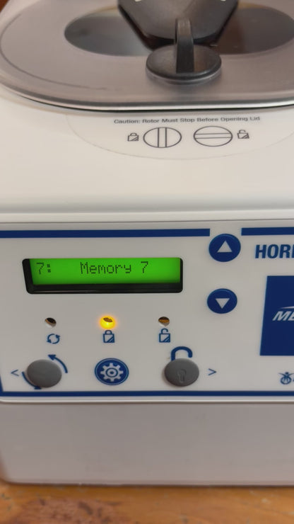 A video demonstration of the Drucker Diagnostics Horizon 6 Flex centrifuge in operation. A gloved hand presses a button on the control panel, changing the digital display from "Memory 7" to a 1-minute cycle at "3500 RPM". As the cycle begins, the lid lock engages (indicated by a lighted lock icon) and the internal chamber illuminates with bright blue LED lighting. The camera looks down through the clear lid at the spinning rotor before panning back to the control panel, where the display toggles between "17